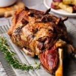 A roasted leg of lamb on a metal plate next to gravy and fresh rosemary.