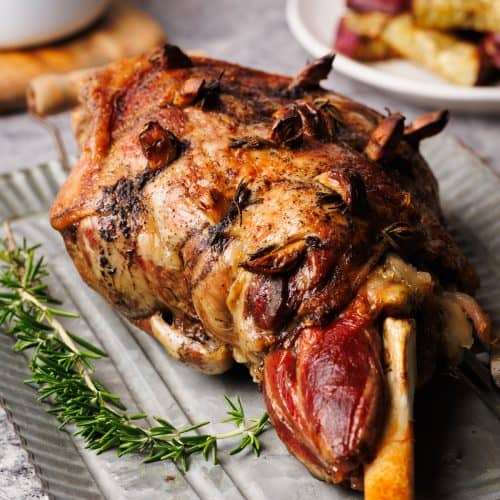 A roasted leg of lamb on a metal plate next to gravy and fresh rosemary.