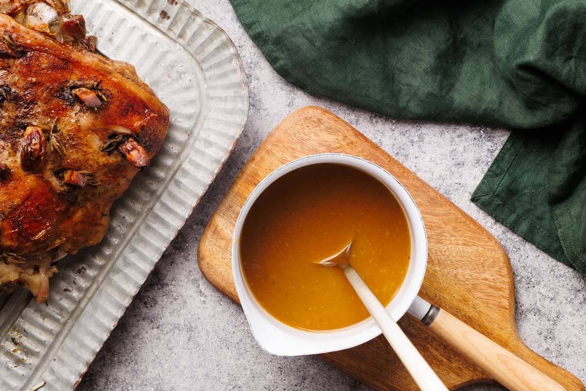 Lamb gravy in a small white saucepan on a wooden board next to roasted leg of lamb.