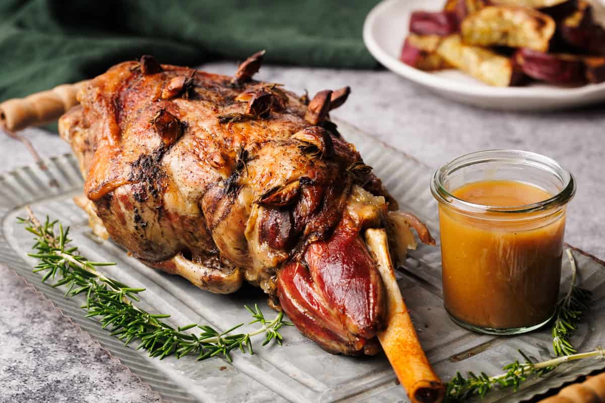 A roasted leg of lamb on a metal plate next to gravy and fresh rosemary.