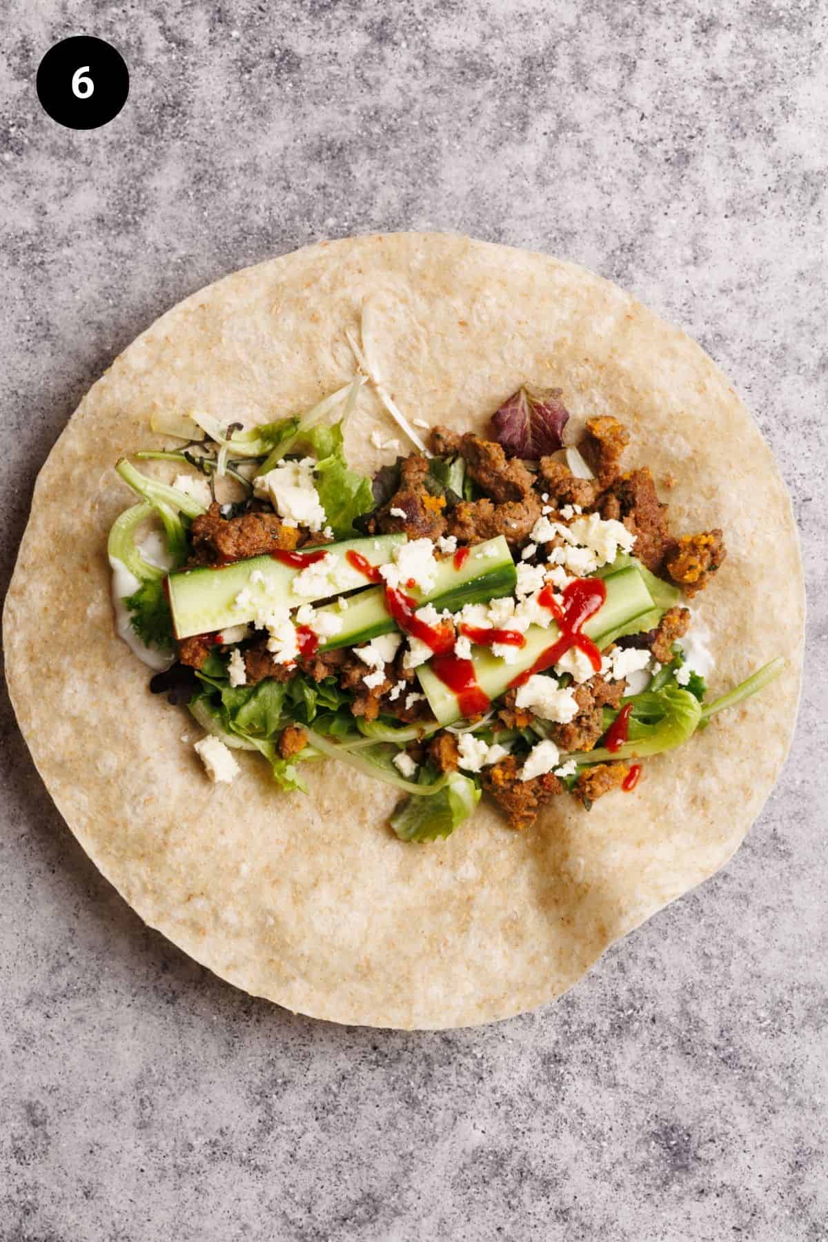 Salad, cucumber, feta, hot sauce, and cooked sausage on a flour tortilla.