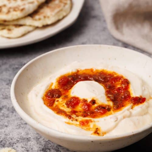 A white bowl filled with harissa yogurt next to a tea towel and flatbread.