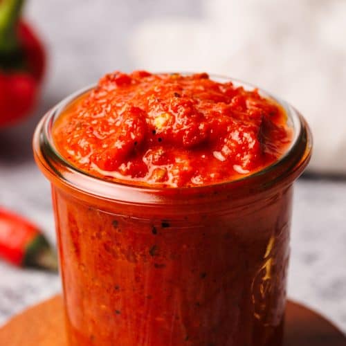 A glass jar filled with harissa paste on a wooden coaster next to chilis.