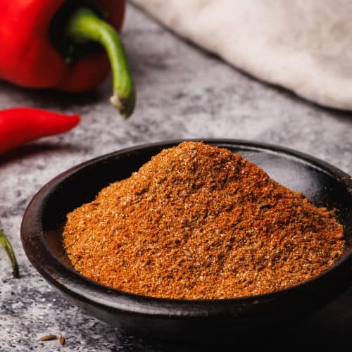Harissa powder in a wooden bowl next to a golden spoon, peppers, and a tea towel.