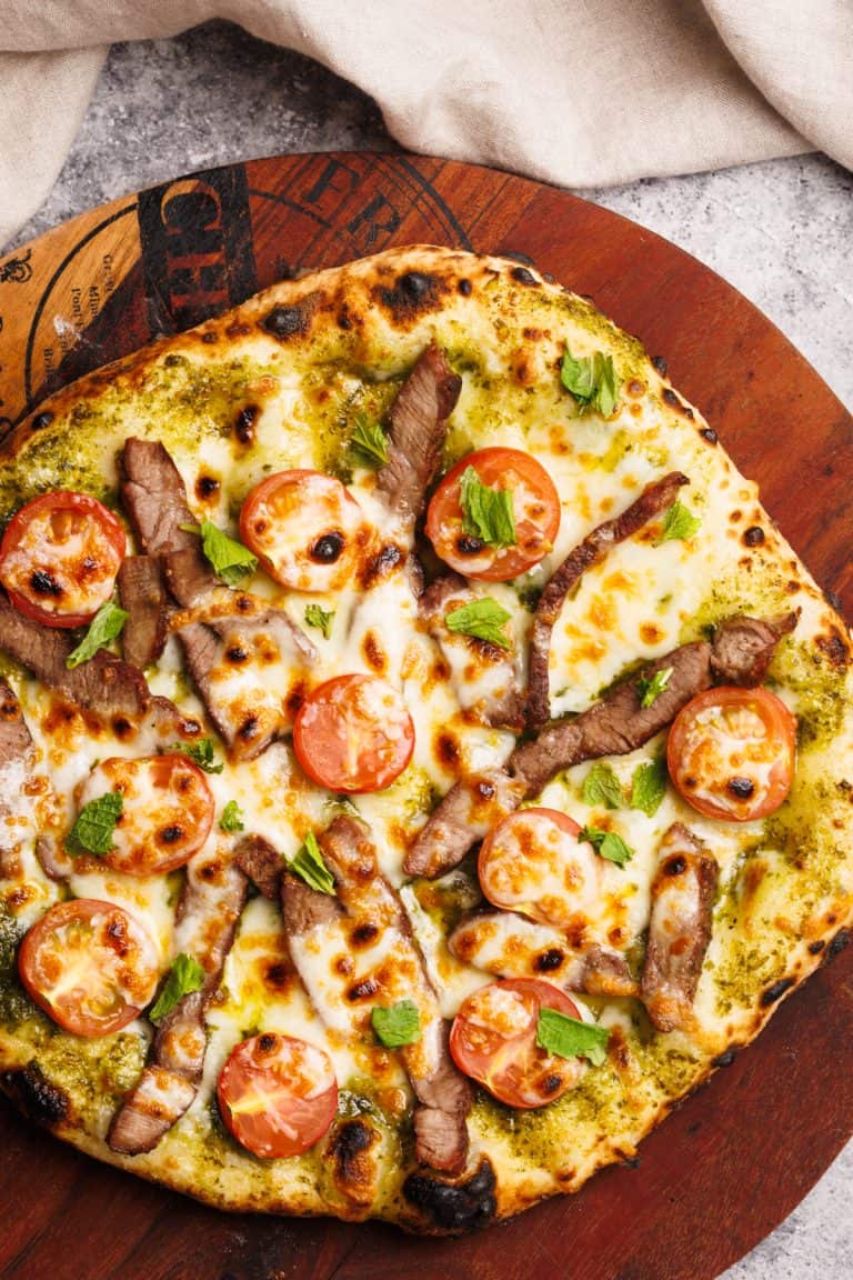 A baked lamb pesto pizza on a wooden board next to a linen napkin.