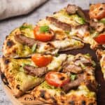 A baked lamb pesto pizza on a wooden board next to a linen napkin.