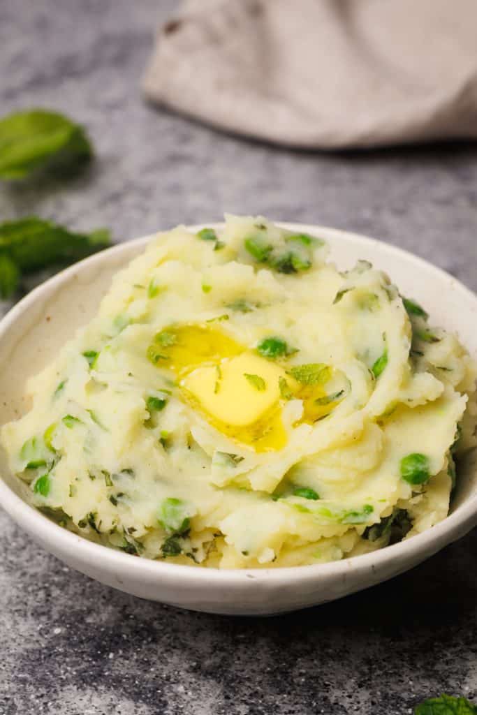 A cream bowl filled with minty mash on a grey background next to fresh mint leaves.