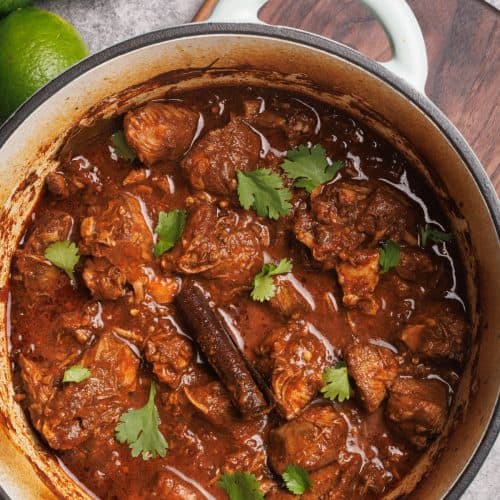 Lamb birria in a Dutch oven, next to fresh coriander, lime and red onion slices.