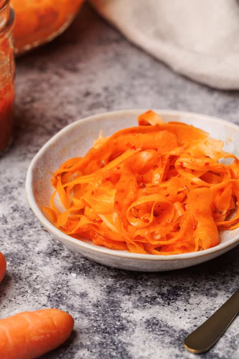 Finished shaved carrot salad in a small bowl next to carrots and a linen napkin.