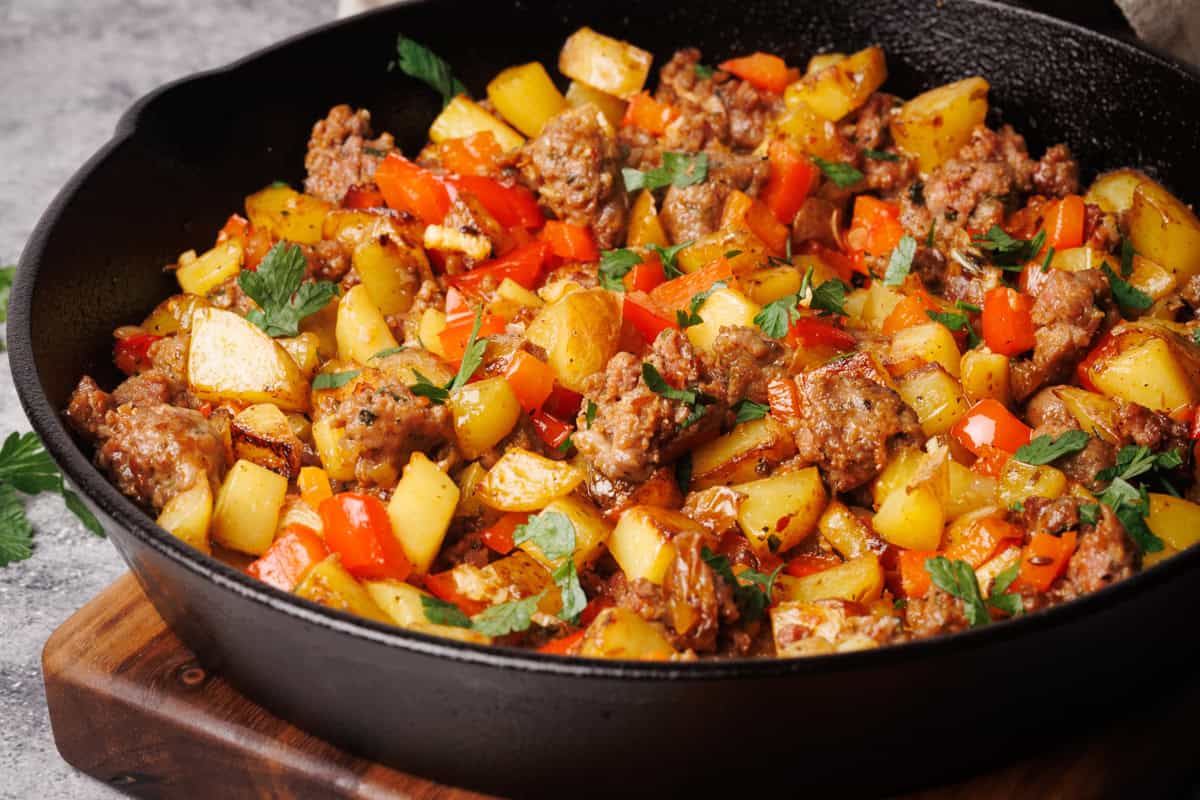 Sausage and potato hash in a cast iron pan next to a linen towel and fresh herbs.