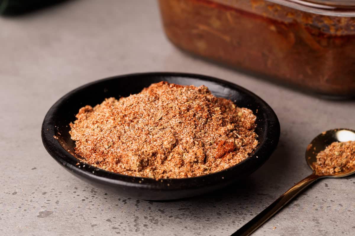 Barbacoa spice mix in a black bowl next to a container of barbacoa.