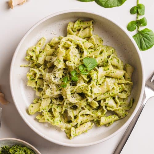 Watercress pesto pasta in a white bowl next to watercress and more pesto.