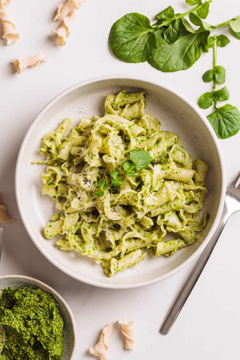Watercress pesto pasta in a white bowl next to watercress and more pesto.