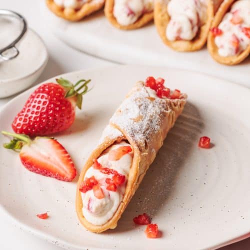 A Strawberry Cannoli on a white plate next to a platter of more.